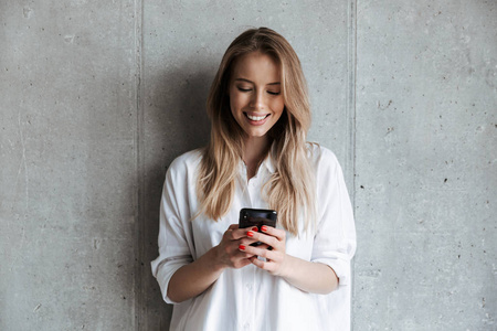 穿着运动衫微笑的年轻女孩站在灰色墙壁背景上使用手机