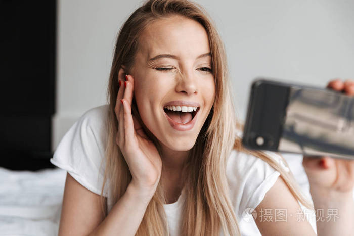 照片的可爱美丽的年轻女子在家室内做自拍手机眨眼照片-正版商用图片0