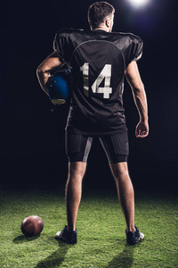 美国橄榄球运动员的后方看法以盔甲站立在绿色草在黑