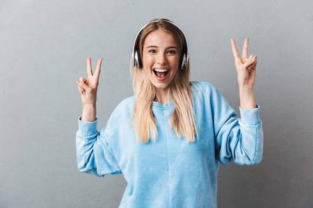 一个开朗的年轻金发女孩的肖像听音乐与耳机和显示和平手势隔离灰色背景