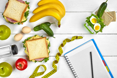 顶部视图规划笔记本与复制空间和健康的早餐。三明治配火腿芝士鸡蛋黄瓜西红柿和瓶装水在白色木桌上
