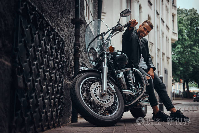 穿着黑色皮夹克和牛仔裤坐在旧欧洲街的复古摩托车的快乐时尚车手