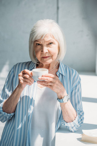 灰色头发妇女的画像与杯子咖啡站立在窗台在家