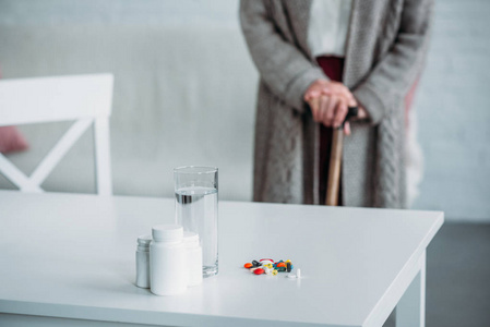 在家里桌上放着药和玻璃杯水的资深妇女有选择地站在房间里
