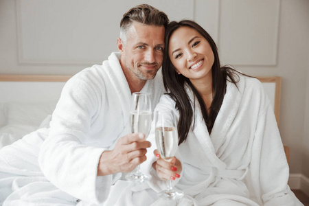 微笑的年轻夫妇穿着浴袍在床上坐着香槟举杯庆祝