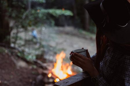 女孩在篝火的背景下喝咖啡。浪漫的气氛。放松的时间露营在山上。活动在假日概念。夜灯