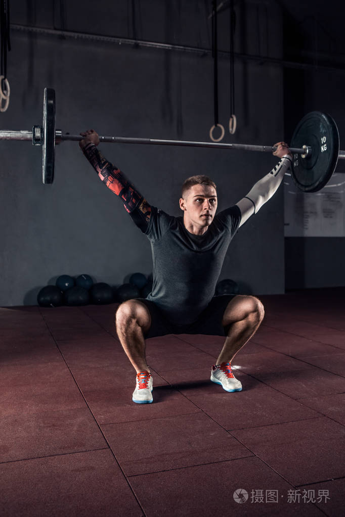 年轻运动员在健身房里用沉重的杠铃在头顶上蹲着.