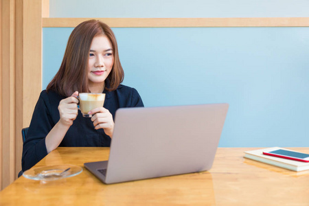 快乐迷人的年轻亚洲商人享受她的咖啡, 而她的笔记本电脑在她的办公室, 良好的现代年轻的工作生活方式的概念