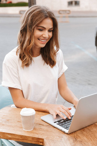夏季日坐在餐桌外的有吸引力的年轻女子的肖像和使用银色笔记本电脑, 同时喝咖啡