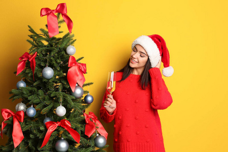 美丽的年轻女子在圣帽与玻璃香槟附近的圣诞树上的颜色背景