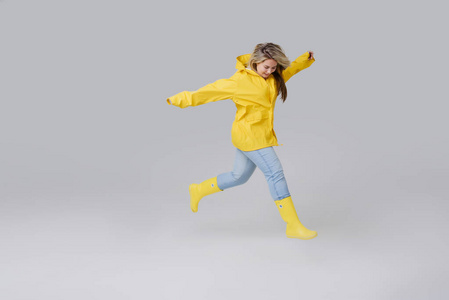 穿着黄色雨衣和黄色橡胶膝靴的年轻女子金发女郎在白色背景上。高兴地步在雨以后跃迁通过水坑