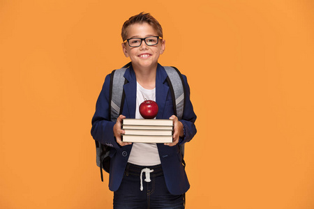 小男孩在眼镜与背包站立在橙色背景, 拿着书和苹果, 微笑