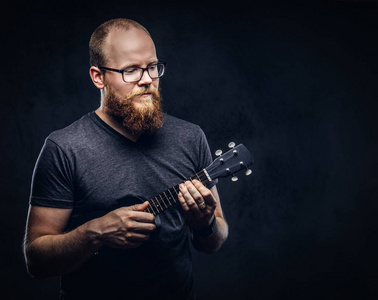 身穿灰色 t恤衫的红胡子男音乐家戴着眼镜, 穿着一双四弦琴。在深色纹理背景下隔离