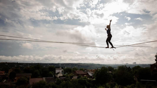 年轻的金发女郎走在 slackline 的绳子上弯曲的膝盖上的房子在树和晴朗的天空的背景