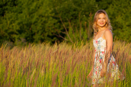美丽的年轻女子站在田野, 绿草和花朵。户外享受大自然。健康微笑的女孩站在高高的草丛中