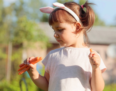 快乐的小女孩吃新鲜和甜胡萝卜。欧洲小女孩与兔耳朵在阳光明媚的夏日在花园里