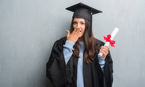 穿灰墙的年轻黑发女子穿着研究生制服持有学位封面与手震惊的错误, 表达恐惧, 害怕沉默, 秘密概念