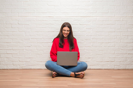 年轻的黑发妇女坐在地板上的白色砖墙使用膝上型电脑与微笑的微笑与自信的笑容显示牙齿