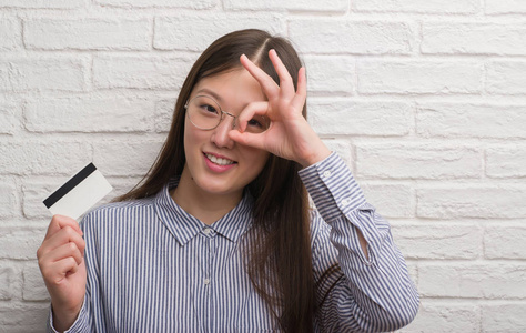 年轻的中国妇女在砖墙持有信用卡与愉快的面孔微笑做 ok 标志用手在眼睛看通过手指