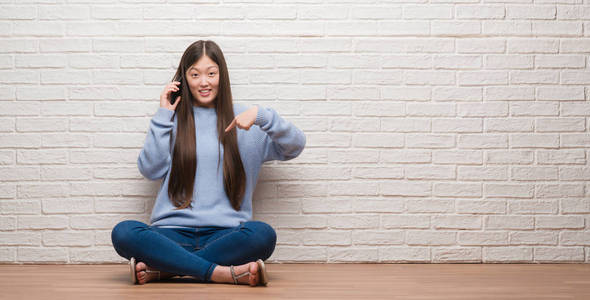 年轻的中国妇女坐在地板上, 用惊讶的脸指着自己的手指在砖墙上