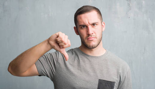 年轻的白种人在灰色垃圾墙壁与愤怒的面孔, 消极标志显示不喜欢与拇指下来, 拒绝概念