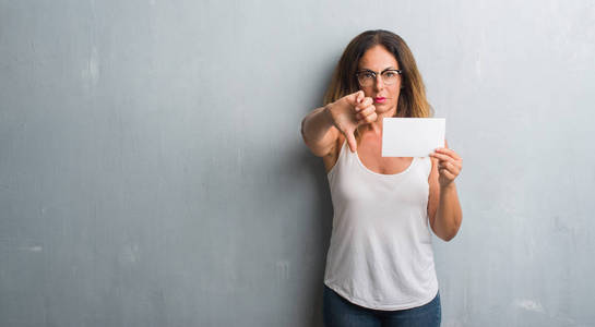 中年拉美裔妇女站在灰色的垃圾墙持有空白卡与愤怒的脸, 消极的迹象显示不喜欢拇指向下, 拒绝概念