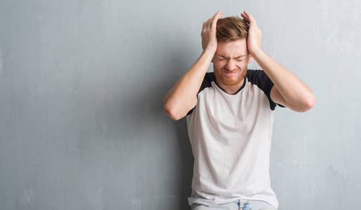 灰色垃圾墙上的年轻红头发的人患有头痛绝望和压力, 因为疼痛和偏头痛。手在头上