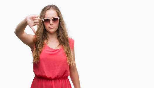 身穿粉红色太阳镜的年轻金发女郎愤怒的脸, 否定的标志显示不喜欢拇指向下, 拒绝概念