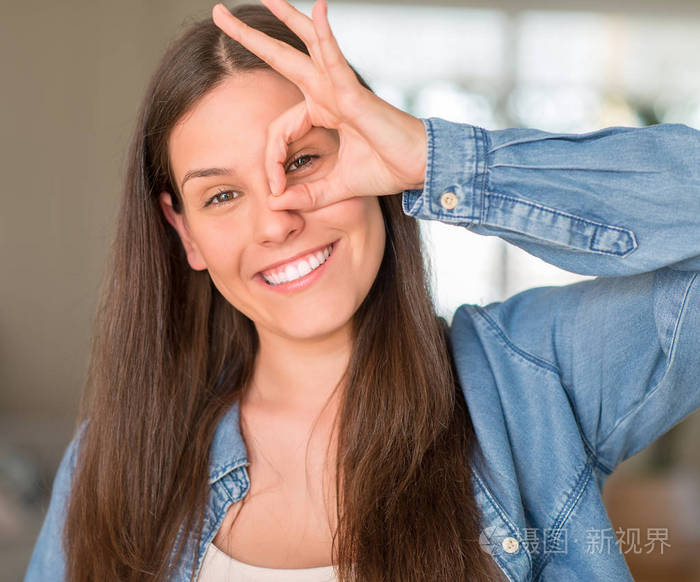 年轻漂亮的女人在家里与快乐的脸微笑做 ok 签名用手在眼睛看通过手指