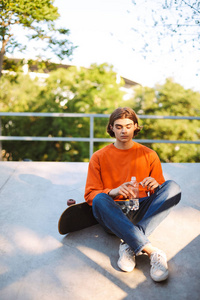 年轻的家伙在橙色的套衫与滑板拿着一瓶水在手上花时间在 skatepark