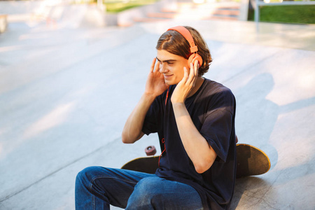 年轻的微笑的家伙在橙色耳机愉快地听音乐与滑板在 skatepark