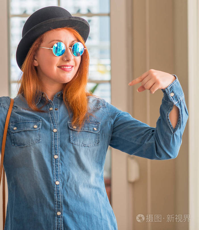 时尚的红发女人戴着圆顶礼帽和太阳镜, 脸上带着微笑, 用手指自豪和