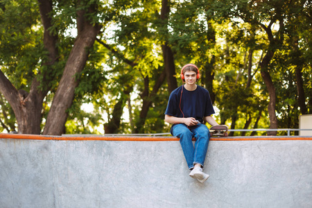 年轻酷的家伙在橙色耳机持有手机和滑板在手花时间在现代 skatepark