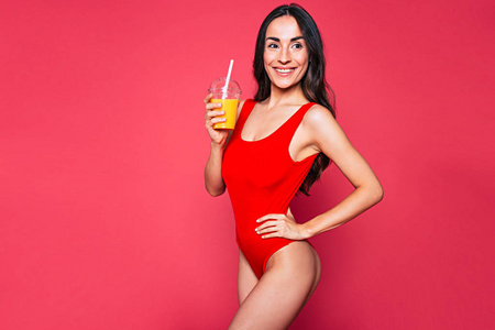 穿着红色泳装的年轻黑发妇女在粉红色的背景上摆出一杯果汁