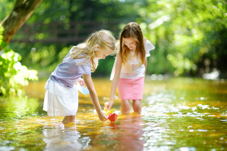 两个小妹妹在温暖晴朗的夏日里在河边玩纸船。孩子们在水里玩得很开心。幼儿暑期活动