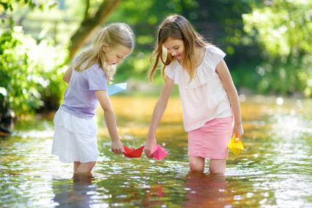 两个小妹妹在温暖晴朗的夏日里在河边玩纸船。孩子们在水里玩得很开心。幼儿暑期活动