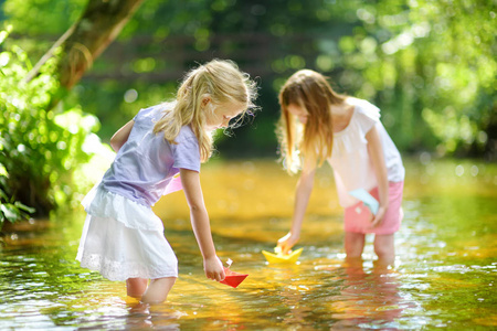 两个小妹妹在温暖晴朗的夏日里在河边玩纸船。孩子们在水里玩得很开心。幼儿暑期活动