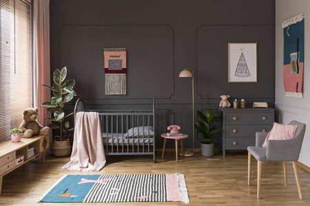 真正的照片, 一个灰色的婴儿床站在粉红色的凳子旁边, 一盏灯和橱柜在灰色婴儿室内部也与扶手椅, 地毯和海报