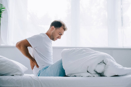 年轻人穿着睡衣坐在床上, 背痛痛苦的侧面观察