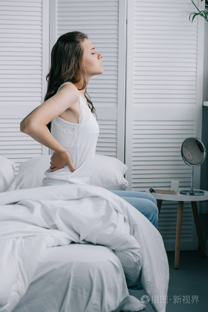 穿着睡衣的年轻女子坐在床上的侧面看着背部疼痛