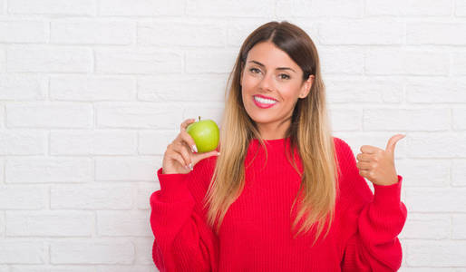 年轻的成年妇女在白色砖墙吃新鲜的绿色苹果指向和显示与拇指到边与愉快的面孔微笑