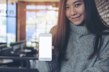 一个亚洲美女的样机图像手持和显示白色手机与空白屏幕在咖啡馆与砖墙背景