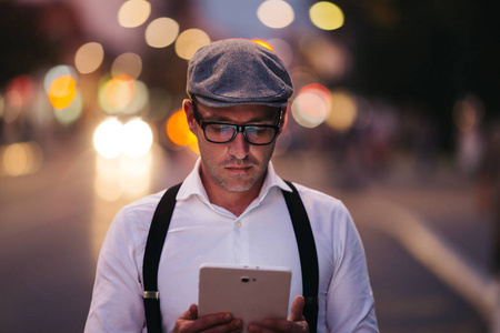 时尚复古穿着帽子, 吊带和眼镜站在城市街道和使用平板电脑的男子。夕阳下的城市灯光