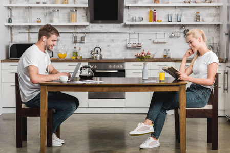 快乐年轻夫妇的侧面视图阅读书和使用笔记本电脑, 而在厨房餐桌上的早餐