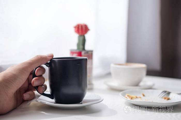 一只手持黑杯热咖啡,白杯,盘子和仙人掌的手特写图片在简约咖啡馆的