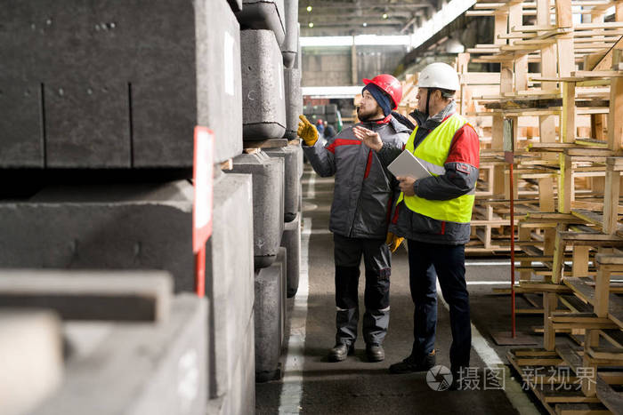 成熟工业工程师的全长肖像穿安全帽检查生产质量与工厂的年轻工人交谈