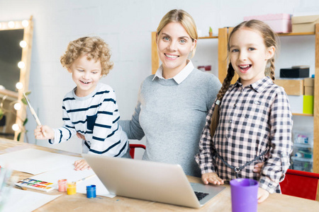 在笔记本电脑上工作的快乐母亲的肖像, 而她的两个孩子画画