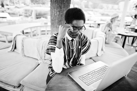 非洲男子穿着传统的衣服和眼镜坐在笔记本电脑后, 在户外咖啡