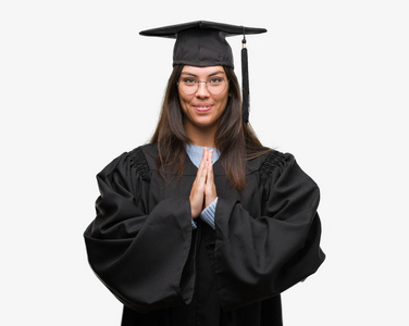 年轻的西班牙裔妇女穿着毕业帽和统一祈祷与双手一起请求原谅微笑自信