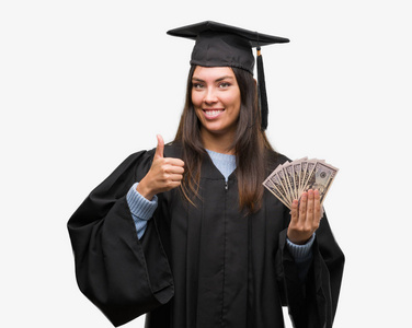 年轻的西班牙裔女子穿着毕业制服持有美元高兴与大微笑做 ok 标志, 拇指与手指, 优秀的标志